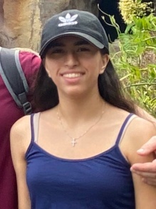 A person wearing a navy tank top and a black baseball cap stands outdoors in front of rocky terrain and greenery. Another person&rsquo;s arm rests lightly on their shoulder from the side of the frame. 