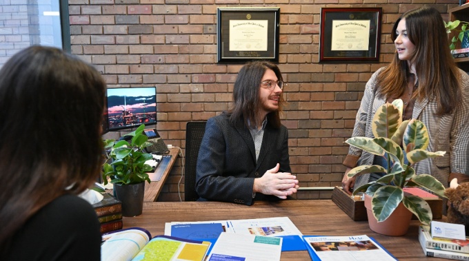 Young man meets with students in his office, admissions materials on his desk. 