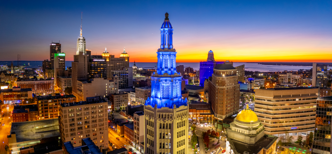 Aerial view of downtown Buffalo at sunset, featuring illuminated skyscrapers and historic buildings, including a blue lit clock tower in the foreground, with Lake Erie visible on the horizon. 