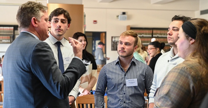 group of young adults standing, listening to person wearing a suit and speaking. 