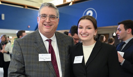 Professionally dressed man and woman standing together, smiling, attending an event in a large open space, with several people standing and talking. 