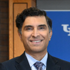A person in a dark suit and patterned tie stands indoors in front of a blue backdrop with white lettering and a wooden panel. 