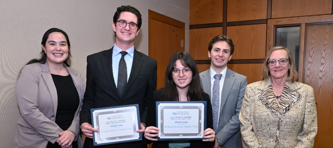 Group photo of five professionally dressed adults standing indoors against a wood-paneled wall. Two people in the center hold framed certificates that read &ldquo;University at Buffalo School of Law, 2025 Charles S. Desmond Moot Court Competition, Winning Team." The group is arranged in a line, with three individuals on the left and two on the right, all facing the camera. The setting appears to be a formal office or event space with neutral lighting and a door visible in the background. 