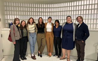Group of adults standing side by side indoors against a glass-block wall, posing for a group photo in business or smart-casual attire. 