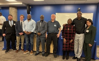 Group of adults standing together at an indoor event space with blue walls, posing for a photo in front of a table and whiteboard. 