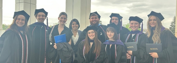 Group of graduates in caps and gowns standing together outdoors, holding diplomas and posing in front of a building with tall columns. 