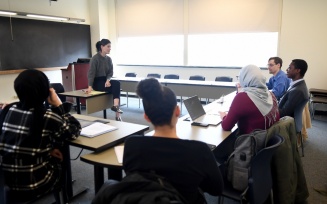 Instructor seated at the front of a classroom leading a discussion, with students seated around tables taking notes and using a laptop. 