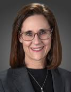 Smiling woman in grey jacket, black shirt, wearing glasses and gold necklace. 