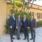 Zoom image: Four people in business attire stand outdoors in front of a Spanish-style building with arches and tall palm trees, posing together while one person holds a plaque labeled &ldquo;Quarterfinalist.&rdquo;