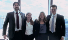 Zoom image: Four people in business attire stand together in front of large windows overlooking a city skyline, posing closely with arms around one another.