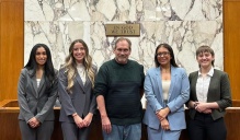 Zoom image: Five people stand side by side at the front of a courtroom, with a marble wall, a state seal, and a sign reading &ldquo;In God We Trust&rdquo; behind them, posing together for a group photo.