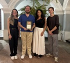 Zoom image: Four people stand indoors beneath an arched ceiling with plants behind them, posing for a photo while one person holds a plaque that reads &ldquo;Second Place.&rdquo;