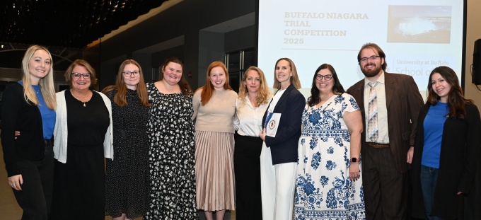 A group of nine people standing together indoors in front of a large projection screen. The screen displays text reading “Buffalo Niagara Trial Competition 2025” along with an image of a scenic waterfront. The individuals are dressed in a mix of business and semi-formal attire, and the setting appears to be a reception or awards event with modern decor and overhead geometric ceiling panels. 