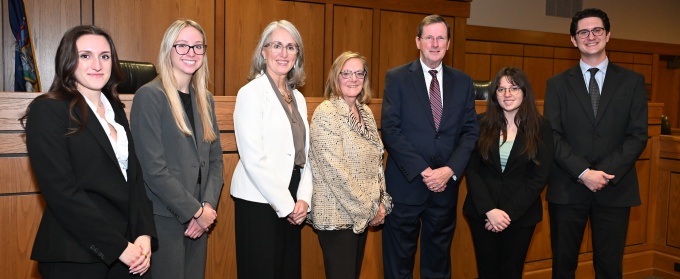 Group of seven people in professional attire posing for a photo in a courtroom in front of the judge’s bench. 