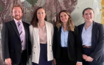 The image shows four individuals standing side by side in front of a textured wall with abstract patterns in muted tones of brown, green, and yellow. All individuals are dressed in professional business attire. 
