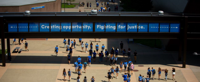 Students wearing blue shirts walk across a campus plaza beneath a pedestrian bridge displaying the message ‘Creating opportunity. Fighting for justice.’ on a bright day. 