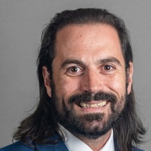 A person wearing a blue suit jacket, white dress shirt, and striped tie, shown against a plain gray background. 