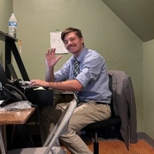 Young man sitting at a desk, smiling and waving while his photo is being taken. 