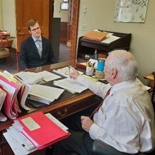 Two men in an office, both sitting across from each other at a desk with lots of papers and folders. 
