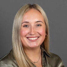 A person with straight, shoulder‑length blonde hair wearing an olive satin button‑down shirt and a small necklace, posed against a plain gray background. 