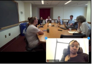 A meeting where people are around a table with one screen window of a woman attending the meeting via Zoom wearing headphones.