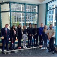 A group of young adults standing next to large windows in an office building, posed for a group photo. 