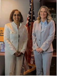 Two women, smiling, standing side by side with US flag between them. 