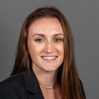 A person with long straight brown hair wearing a black blazer over a white top and a thin layered necklace, posed against a plain gray background. 