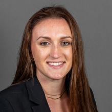 A person with long straight brown hair wearing a black blazer over a white top and a thin layered necklace, posed against a plain gray background. 