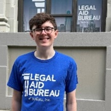 Young man wearing a blue t-shirt that says Legal Aid Bureau, standing in front of a building. 