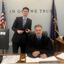 Young man standing next to man wearing judges robes, sitting at a court bench. 