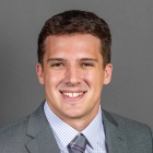 A person wearing a gray suit jacket with a lapel pin, a light checkered dress shirt, and a gray plaid tie, posed against a plain gray background. 
