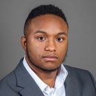 A person wearing a gray suit jacket over a white dress shirt, posed against a plain gray background. 