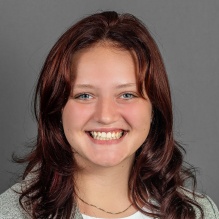 A person with shoulder‑length wavy dark auburn hair wearing a light gray blazer over a white blouse, posed against a plain gray background.
