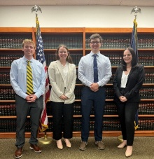 Four young professional adults, lined up for photo inside a room with bookshelves and flags.