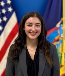 young woman smiling, US and NY state flags behind her.