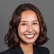 A person with short wavy dark brown hair wearing a black blazer over a bright blue top, posed against a plain gray background. 
