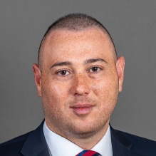 A person wearing a dark suit jacket, white dress shirt, and a red‑and‑blue striped tie, posed against a plain gray background. 