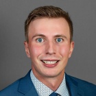 A person with short light brown hair wearing a blue suit jacket, a patterned light blue dress shirt, and a navy textured tie, posed against a plain gray background. 