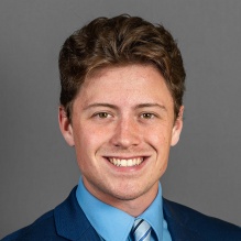 A person with short wavy brown hair wearing a blue suit jacket, a light blue dress shirt, and a striped blue tie, posed against a plain gray background. 