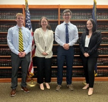 Four young professional adults lined up for a photo in a room with bookshelves and flags. 