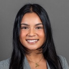 A person with long dark hair wearing a gray plaid blazer over a black top and a gold necklace with a pendant, posed against a plain gray background. 