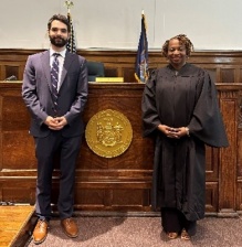 Young professional man standing next to woman dressed in judges robe, standing in front of a judges bench with gold seal between them.