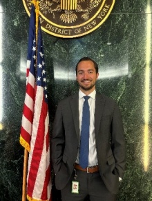 Young professional man standing against a green marbled wall next to a US flag and under a gold emblam. 