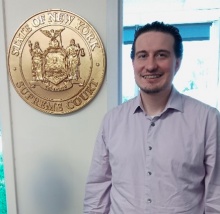 Young man standingin front of a window, next to a gold emblam that says State of New York Supreme Court.