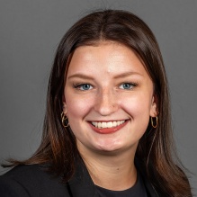 A person with shoulder‑length brown hair wearing gold hoop earrings, a black blazer, and a dark top, posed against a plain gray background.