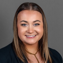 A person with straight shoulder length brown hair wearing a black blazer over a black top and a gold lariat style necklace, posed against a plain gray background. 