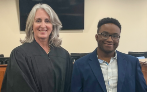 Two people standing together in a courtroom, with a judge’s bench and seating visible in the background.