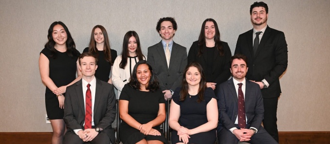 Group portrait of ten people in formal business attire arranged in two rows, with four seated on chairs in the front row and six standing behind them. The photo is taken indoors against a neutral wall with patterned carpet flooring, and all individuals are posed facing the camera. 