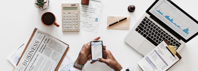 photo of hands over a white desk with a newspaper, laptop, and phone. 